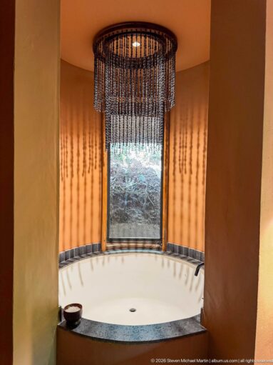 Recessed soaking tub in an ochre alcove beneath a beaded pendant light, with a narrow window facing vegetation.