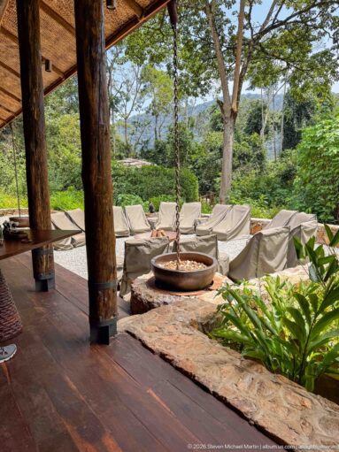 Timber terrace with heavy posts overlooking a hanging fire bowl, covered lounge chairs, trees and distant hills.