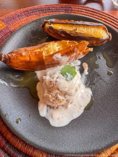 Two roasted plantain- or banana-like halves beside a creamy garnished accompaniment on a dark ceramic plate.