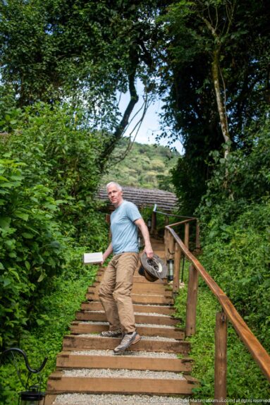 Man descending broad outdoor steps with timber rails through dense vegetation, carrying personal items beside the lodge.