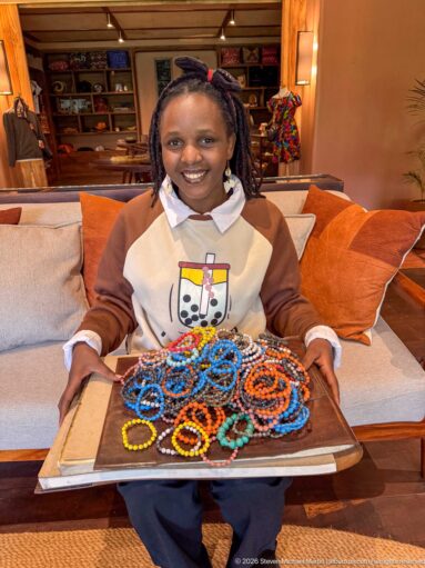 Smiling seated woman holding a tray covered with colorful beaded bracelets and necklaces inside the lodge.