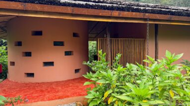 Curved ochre lodge wall with small rectangular openings beneath a thatched roof, bordered by green foliage.