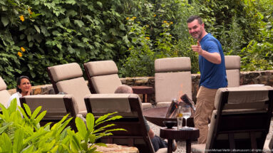 Standing man and seated guest beside a small fire pit, cushioned chairs and drinks in a lush forest setting.