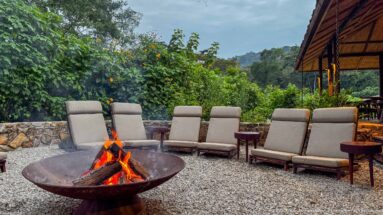 Flaming metal fire bowl surrounded by cushioned lounge chairs on gravel with flowering shrubs and forested hills behind.