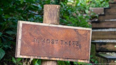 Weathered metal sign reading “ALMOST THERE” mounted on a wooden post beside outdoor steps and green vegetation.