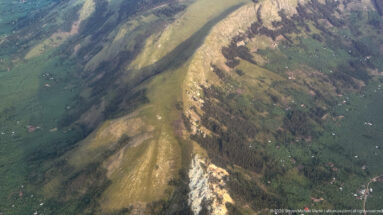 Steep green escarpment with pale exposed rock, cultivated plots, woodland, and scattered houses.