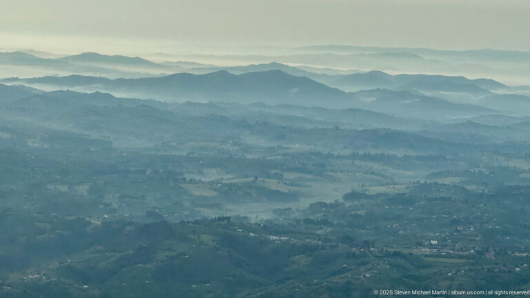 Layered blue-gray mountain ridges with cultivated slopes and scattered settlement in western Uganda.