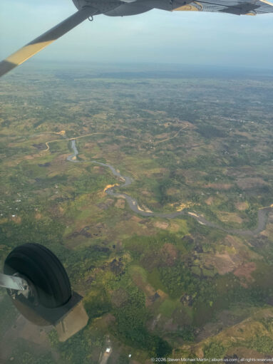 Aircraft wheel and wing frame a winding river, wetlands, and farmland below.