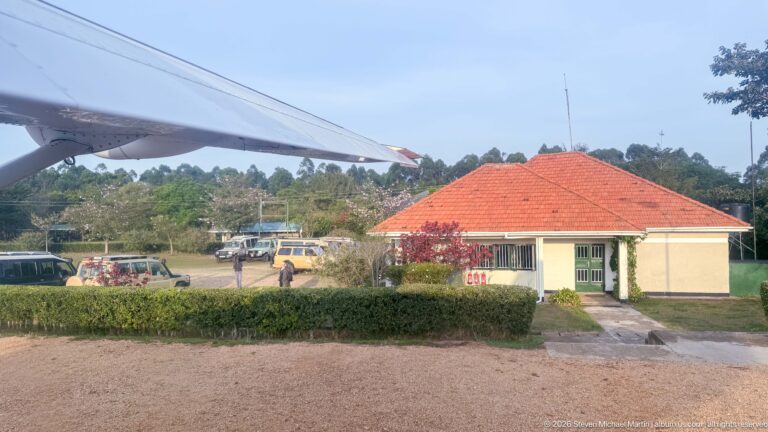 Red-roofed Kihihi terminal and parked vehicles seen beneath the wing of a departing light aircraft.