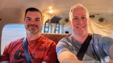 Two smiling male passengers wearing shoulder belts inside the compact cabin of a regional light aircraft.