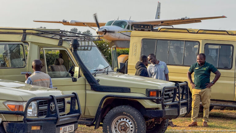 Men stand beside safari vehicles with a high-wing light aircraft parked immediately behind them at Kihihi.
