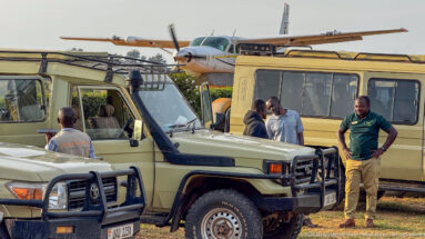 Men stand beside safari vehicles with a high-wing light aircraft parked immediately behind them at Kihihi.