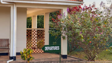 Covered veranda at Kihihi with green Departures sign, flowering shrubs, white columns, and a lattice screen.