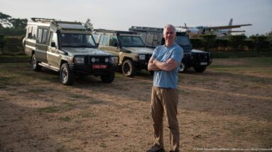 Traveler with crossed arms stands beside parked four-wheel-drive safari vehicles with part of a small aircraft behind.