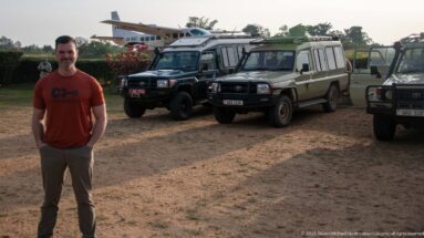Traveler stands before several four-wheel-drive safari vehicles with a small aircraft visible in the background.