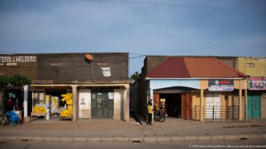 Open shops with stacked goods and a red-roofed financial-service storefront line a paved road in Kihihi.