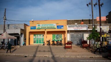 Kyamuhunga Peoples SACCO Kihihi Branch stands among neighboring roadside businesses, motorcycles and pedestrians.