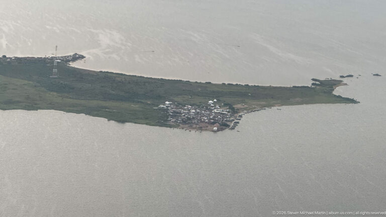 Compact lakeside settlement clustered at the end of a narrow green peninsula surrounded by gray water.