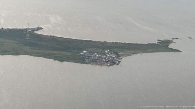 Compact lakeside settlement clustered at the end of a narrow green peninsula surrounded by gray water.