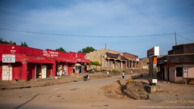 Low shops, including a red Airtel-branded block, line a broad unpaved street with pedestrians and a motorcycle.