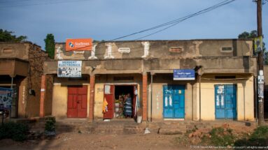 Weathered concrete shops with red and blue doors, including a signed Herbert Electronics storefront, face the road in Kihihi.