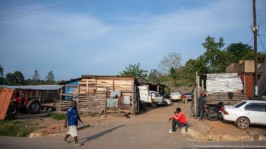 Pedestrians, parked cars, motorcycles and a tractor line an unpaved lane between timber and metal workshops.