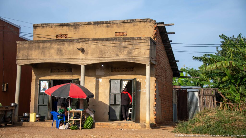 Small concrete and brick storefront with an umbrella-shaded table, blue chair and banana plants beside it.