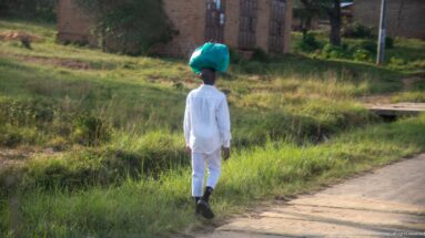 Person in white walks away along a roadside carrying a large turquoise-wrapped load on the head.