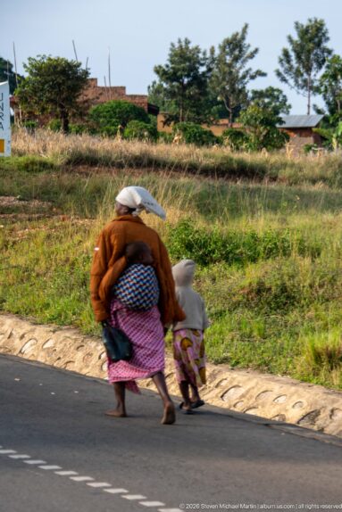Adult carrying a bundled load walks hand in hand with a small child along the edge of a paved road.