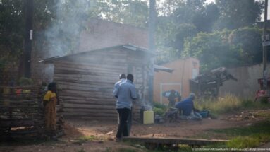Several people stand and sit beside a rough timber structure as smoke drifts through a Kihihi neighborhood.