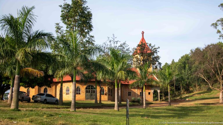 Brick church with arched windows, red roof and pointed tower behind palm trees on a grassy compound.