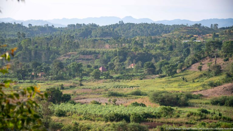 Patchwork fields, scattered houses, wooded hills and a distant mountain ridge in the rural landscape around Kihihi.