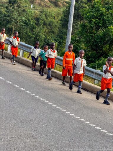 Young schoolchildren in orange and pale uniforms walking along a paved road beside a metal guardrail.