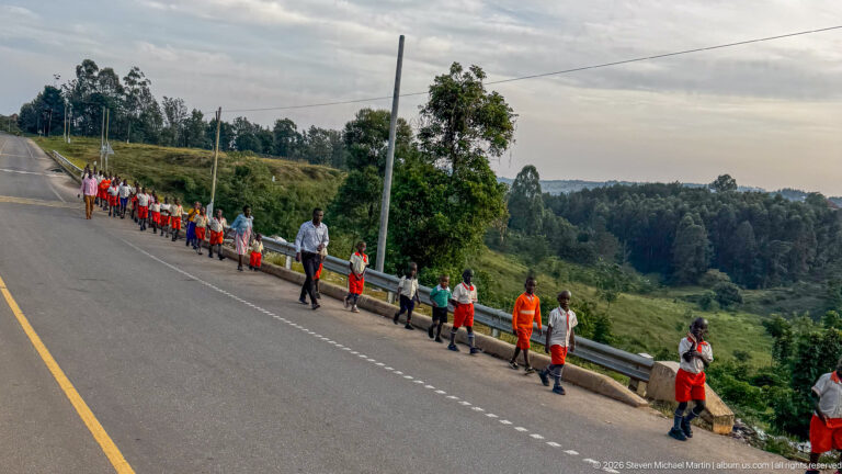 Line of schoolchildren in orange, white, and green uniforms walking beside a paved road with adults in hilly countryside.