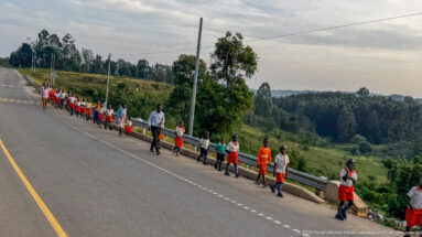 Line of schoolchildren in orange, white, and green uniforms walking beside a paved road with adults in hilly countryside.