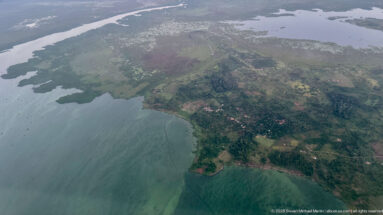 Settled green peninsula with fields and buildings projecting into pale blue-green lake water.