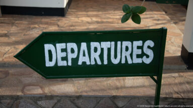 Dark green arrow-shaped sign reading DEPARTURES beside a stone-paved area at the airfield.