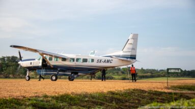 Aerolink Uganda high-wing turboprop parked on a gravel airstrip with ground staff and an ARRIVALS sign nearby.