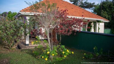 Single-story pale building with red tile roof, shaded veranda, barred windows, and flowering garden.