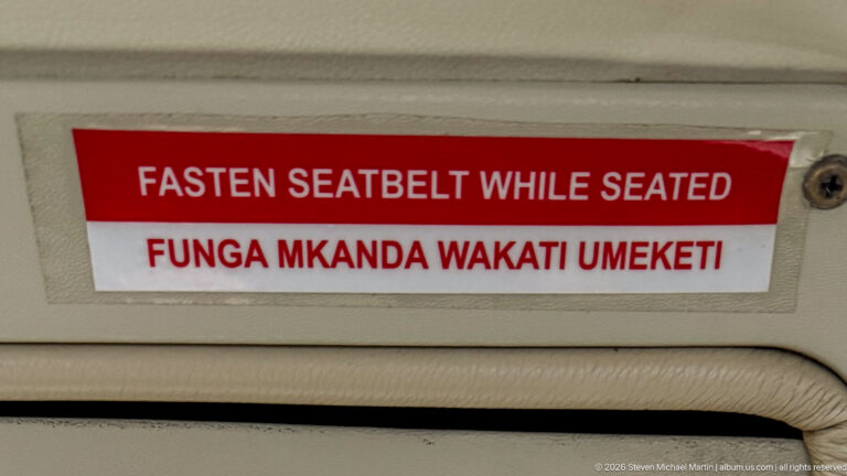 Red-and-white aircraft placard reading Fasten Seatbelt While Seated with the same instruction in Swahili below.