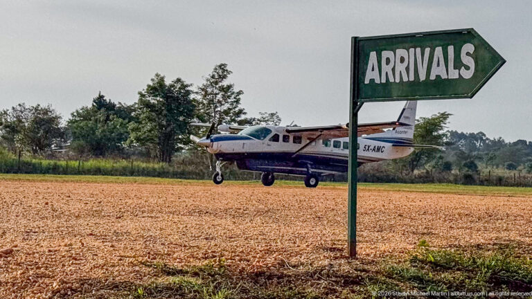 Green ARRIVALS arrow sign beside a gravel airstrip with a small high-wing aircraft in the background.