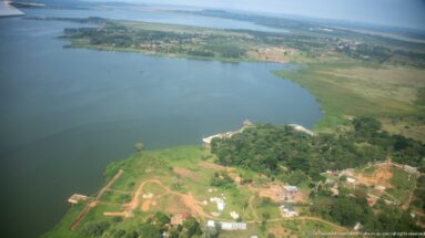 Oblique aerial view of a large lake bordered by green fields, scattered buildings, trees, and a small shoreline landing.