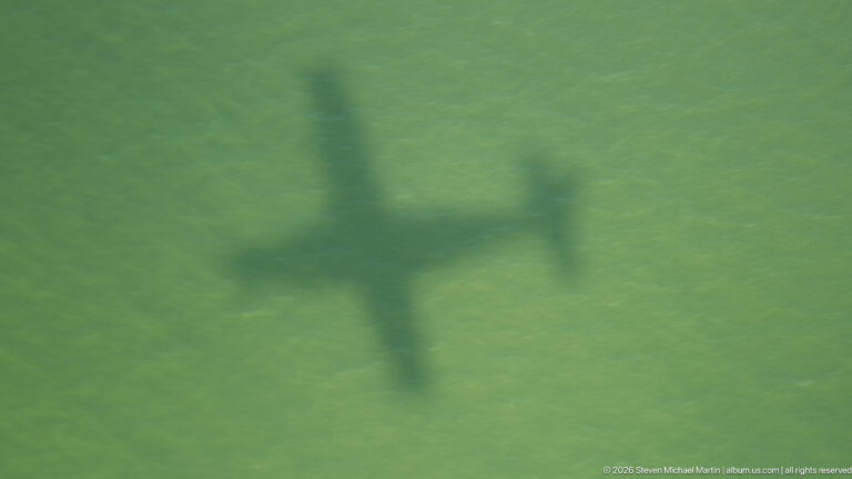 Aircraft-shaped shadow cast across a uniform green landscape, with wings, fuselage, and tail clearly outlined.