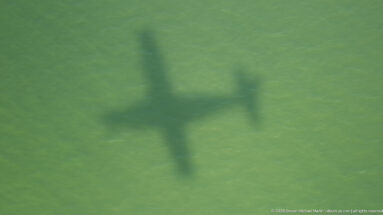 Aircraft-shaped shadow cast across a uniform green landscape, with wings, fuselage, and tail clearly outlined.