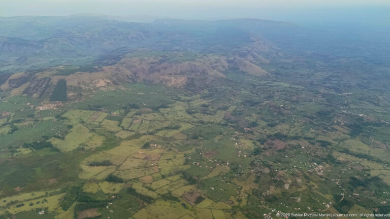 Patchwork fields, villages, tree belts, and roads extending toward low ridges in western Uganda.