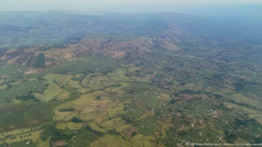 Patchwork fields, villages, tree belts, and roads extending toward low ridges in western Uganda.