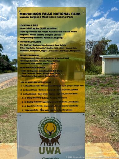 Yellow Murchison Falls National Park visitor information board with regulations and Uganda Wildlife Authority emblem beside a road.