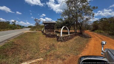 Sambiya River Lodge entrance marker beside a paved highway and red dirt track, with part of a vehicle in foreground.