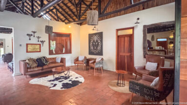 Angled lodge lounge with sofa and chairs, hide rug, wooden doors, mirrors, lion artwork, and exposed roof.