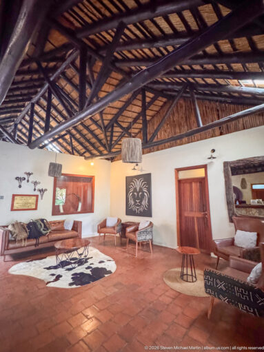 Wide lodge sitting room with scattered seating, rugs, lion wall art, terracotta tiles, and an exposed timber roof.